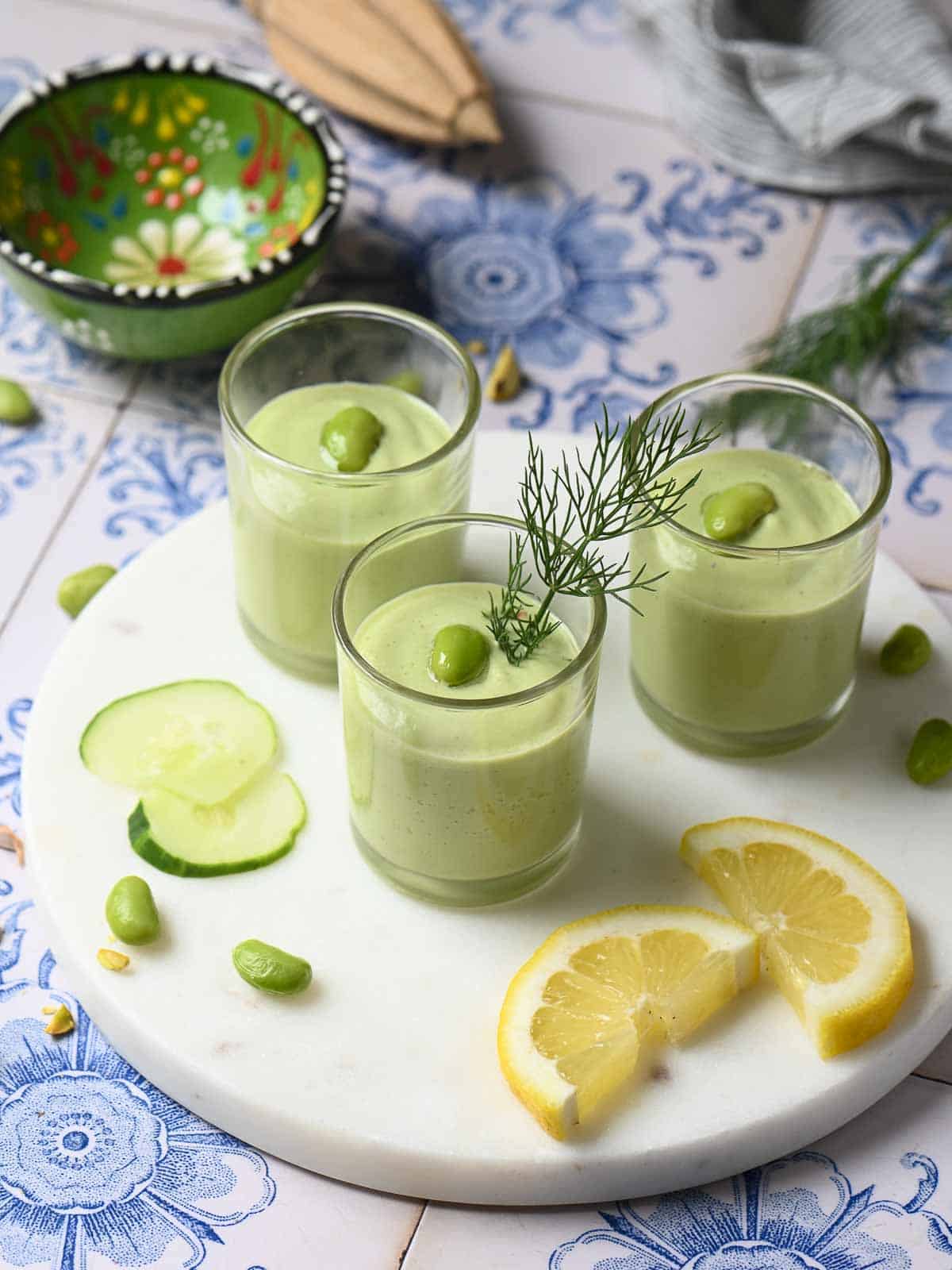 Three cucumber soups in clear shot glasses with various toppings scattered around them.