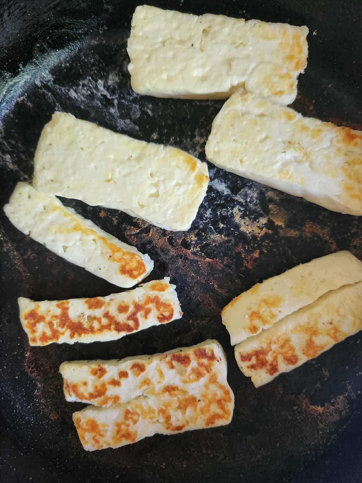 Slices of halloumi fried in a black skillet.