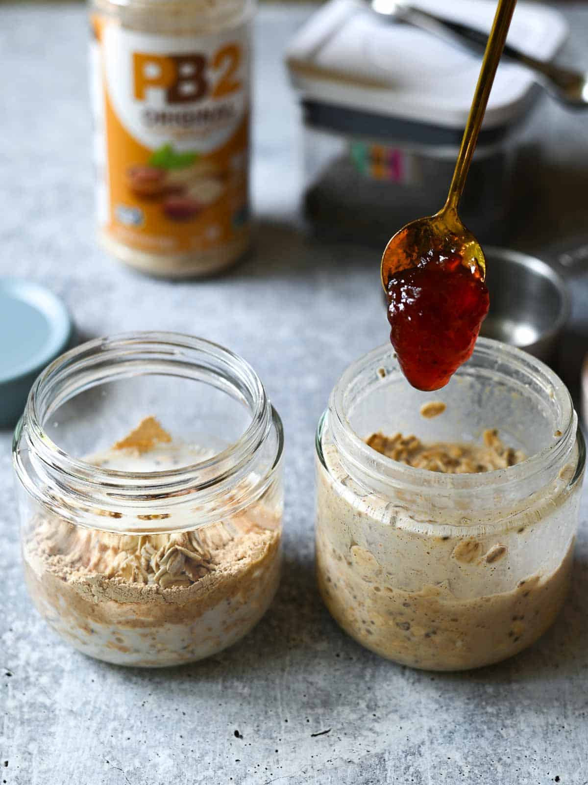 Jam being spooned into a jar of overnight oats on a concrete surface.
