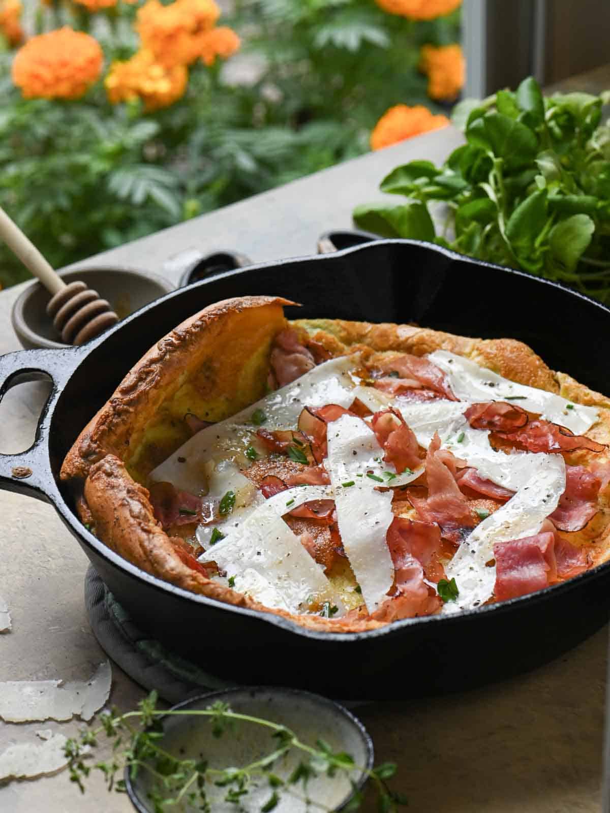 A Dutch baby pancake in a black skillet with greens and orange flowers in the background.