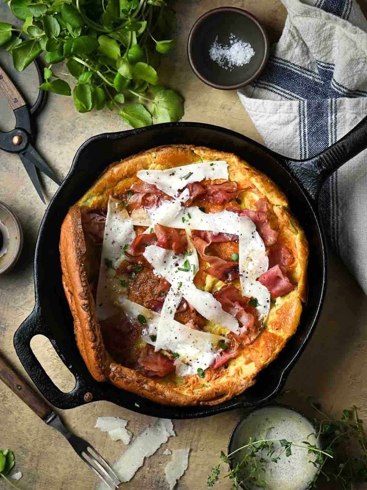 Overhead of a savory Dutch baby in a skillet with herbs and a towel next to it.