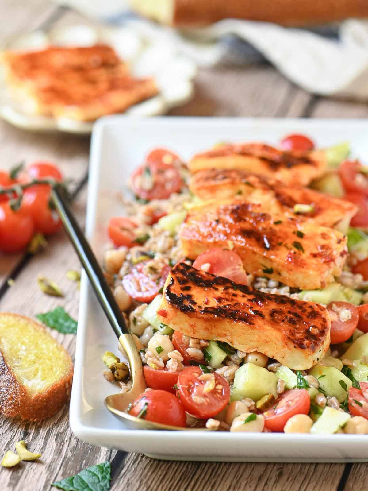 Grilled halloumi salad on a rectangular white plate with tomatoes and bread to the side.