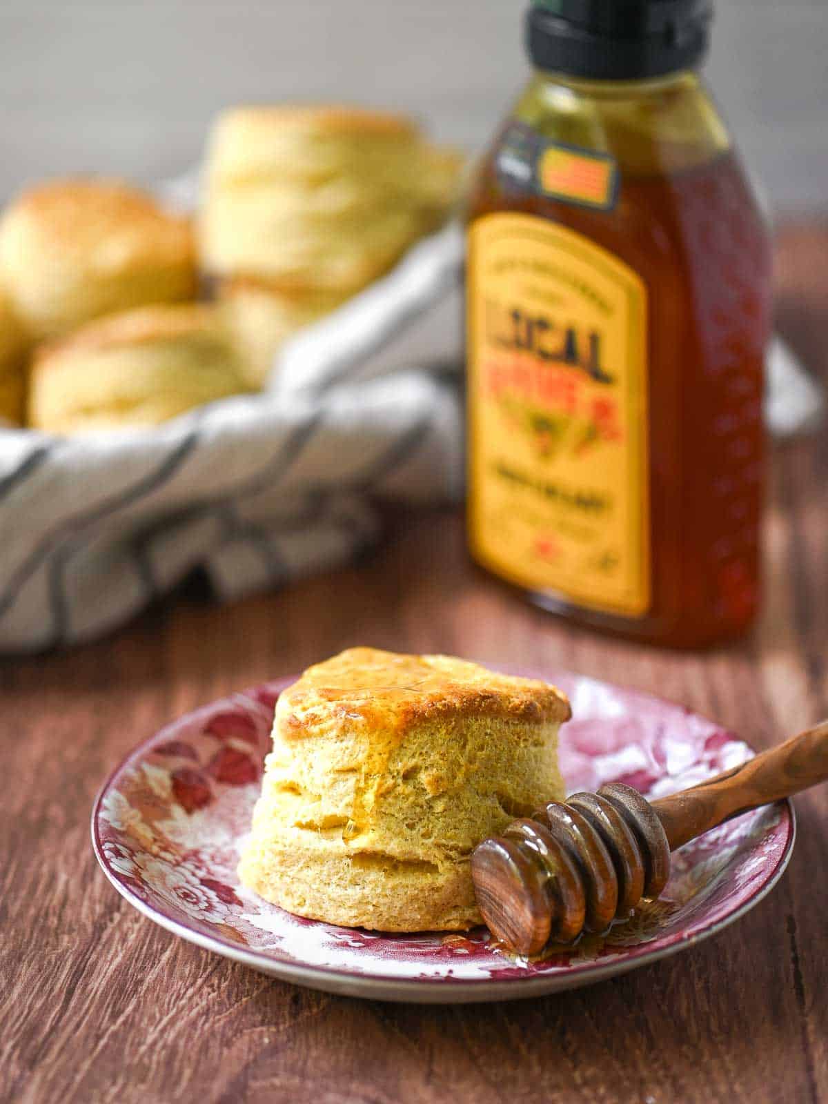A biscuit on a flowered plate with honey dripping down from it and a bottle of honey in the background.