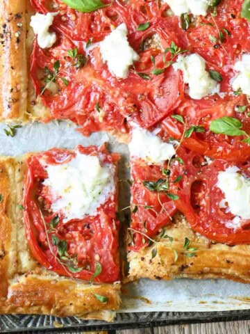 A puffed pastry tomato tart on a metal pan with a slice cut out.