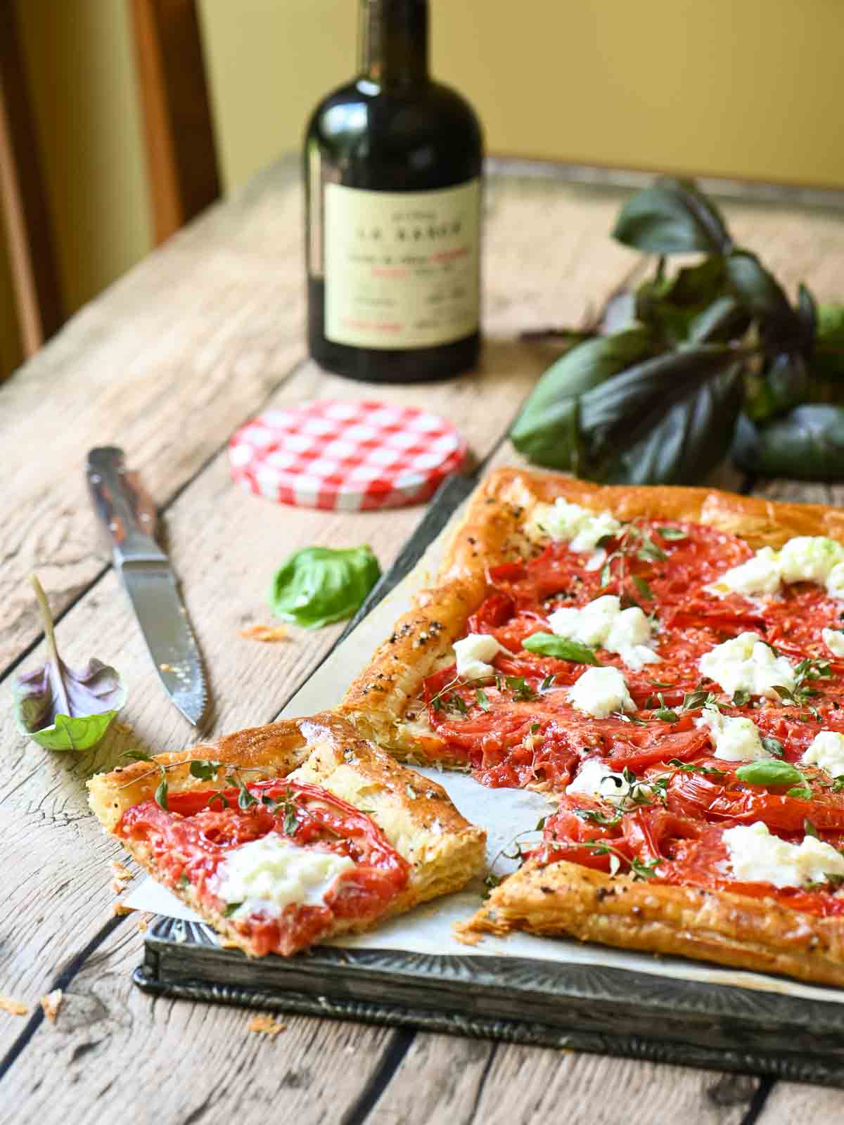 A table view of a tomato tart cut up with fresh basil around it and a bottle of oil.