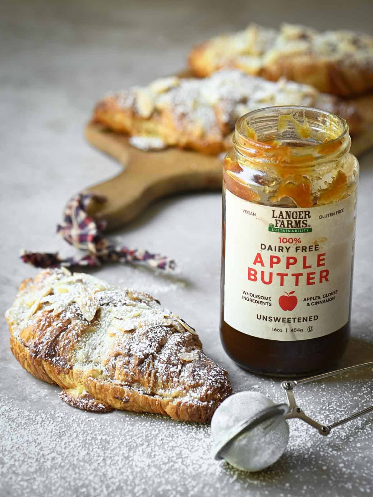An almond croissant placed to the left of a jar of apple butter on a beige surface.