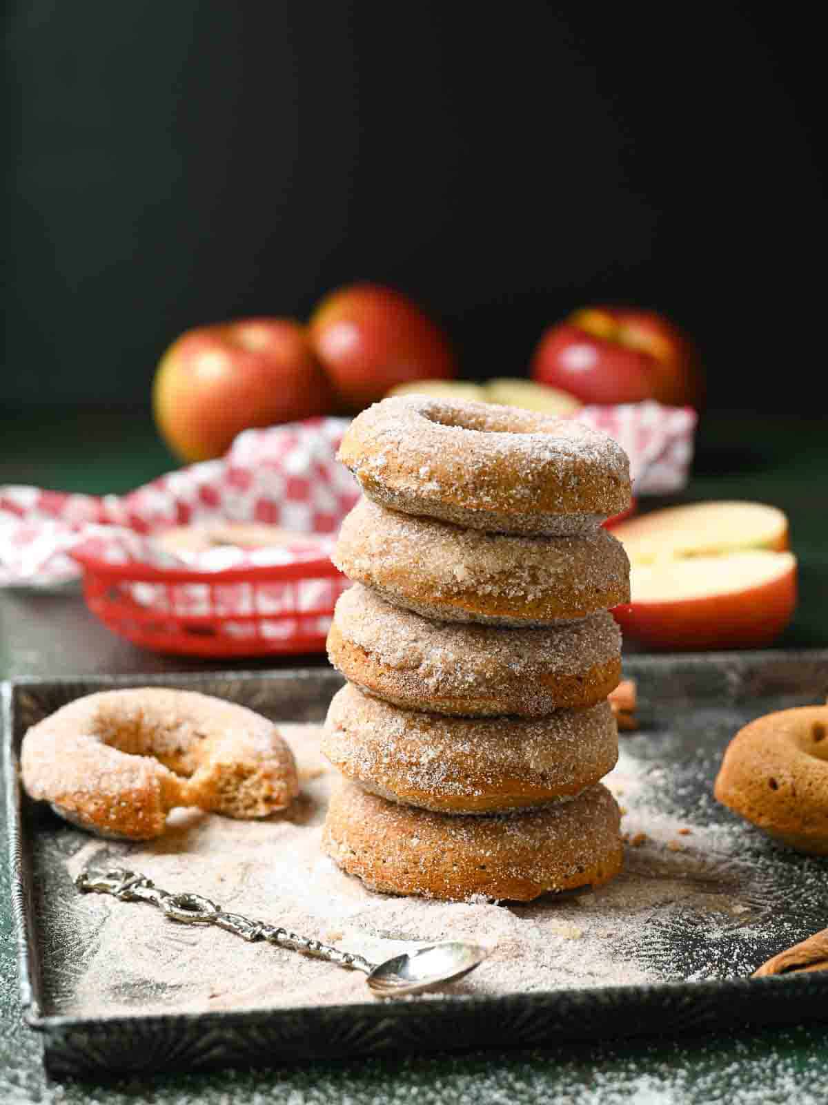 A stack of apple donuts on a metal tray with apples in the background.