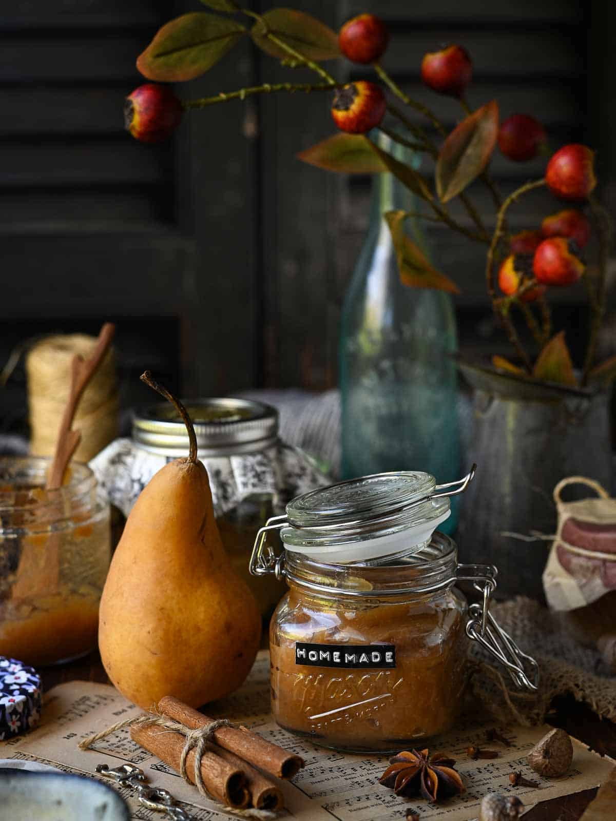 Pear butter in a clear Mason jar with a whole pear next to it and a vase of orange branches in the back.